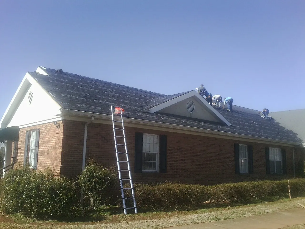 Crew tearing shingles off a brick ranch roof with a ladder set — Emergency Roof Repair work by our crew in Indian Lake Estates