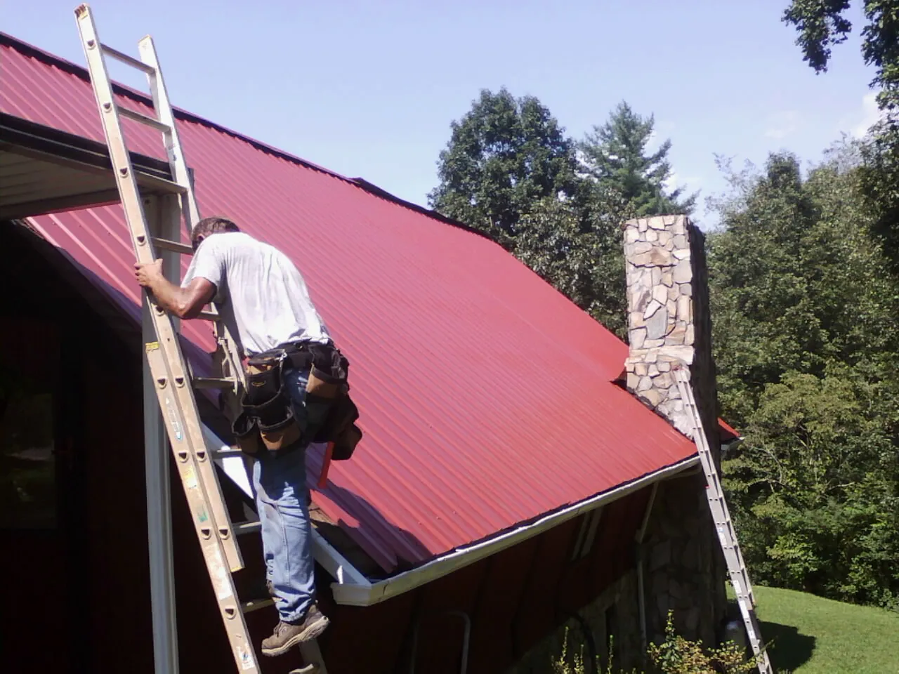 Roofer on a ladder beside a red metal roof and stone chimney — Soffit & Fascia Repair by Roofing Brothers in Indian Lake Estates